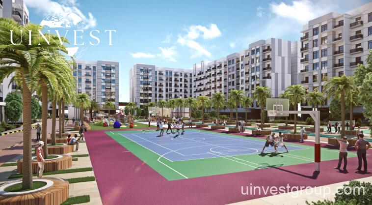 Exterior rendering of Lawnz by Danube Properties in Dubai International City, showing four eight-storey, country-themed freehold apartment buildings with pastel facades, geometric motifs, private balconies, surrounded by landscaped gardens, a community swimming pool, outdoor sports courts, and palm trees under a bright blue sky.