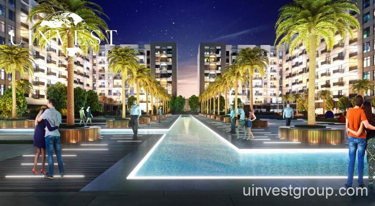 Exterior rendering of Lawnz by Danube Properties in Dubai International City, showing four eight-storey, country-themed freehold apartment buildings with pastel facades, geometric motifs, private balconies, surrounded by landscaped gardens, a community swimming pool, outdoor sports courts, and palm trees under a bright blue sky.