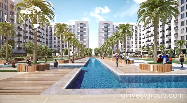 Exterior rendering of Lawnz by Danube Properties in Dubai International City, showing four eight-storey, country-themed freehold apartment buildings with pastel facades, geometric motifs, private balconies, surrounded by landscaped gardens, a community swimming pool, outdoor sports courts, and palm trees under a bright blue sky.