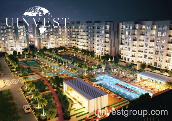 Exterior rendering of Lawnz by Danube Properties in Dubai International City, showing four eight-storey, country-themed freehold apartment buildings with pastel facades, geometric motifs, private balconies, surrounded by landscaped gardens, a community swimming pool, outdoor sports courts, and palm trees under a bright blue sky.
