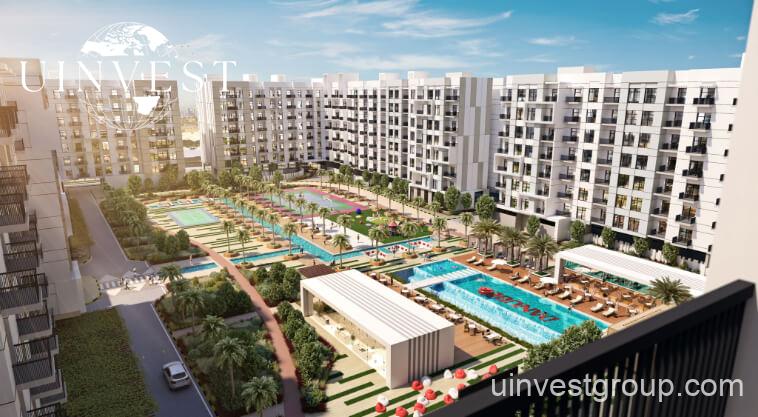 Exterior rendering of Lawnz by Danube Properties in Dubai International City, showing four eight-storey, country-themed freehold apartment buildings with pastel facades, geometric motifs, private balconies, surrounded by landscaped gardens, a community swimming pool, outdoor sports courts, and palm trees under a bright blue sky.