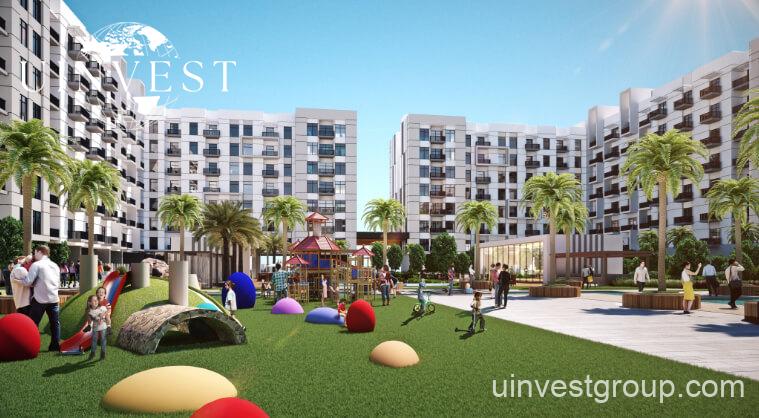Exterior rendering of Lawnz by Danube Properties in Dubai International City, showing four eight-storey, country-themed freehold apartment buildings with pastel facades, geometric motifs, private balconies, surrounded by landscaped gardens, a community swimming pool, outdoor sports courts, and palm trees under a bright blue sky.