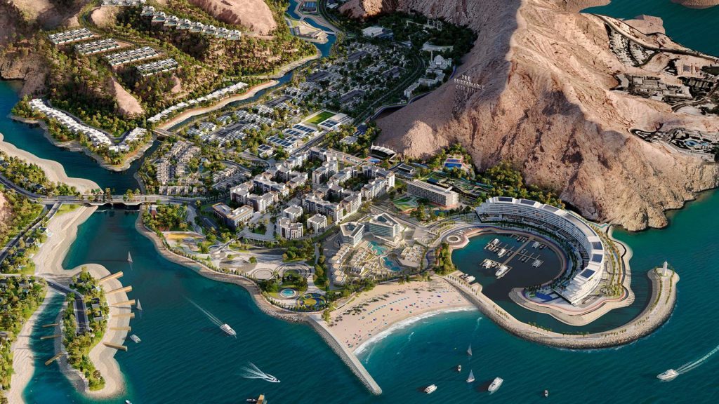 Aerial view of The Sustainable City Yiti on the Gulf of Oman coast, showing the marina, beach, The Arc Residences and the surrounding mountains.
