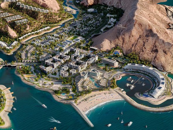 Aerial view of The Sustainable City Yiti on the Gulf of Oman coast, showing the marina, beach, The Arc Residences and the surrounding mountains.