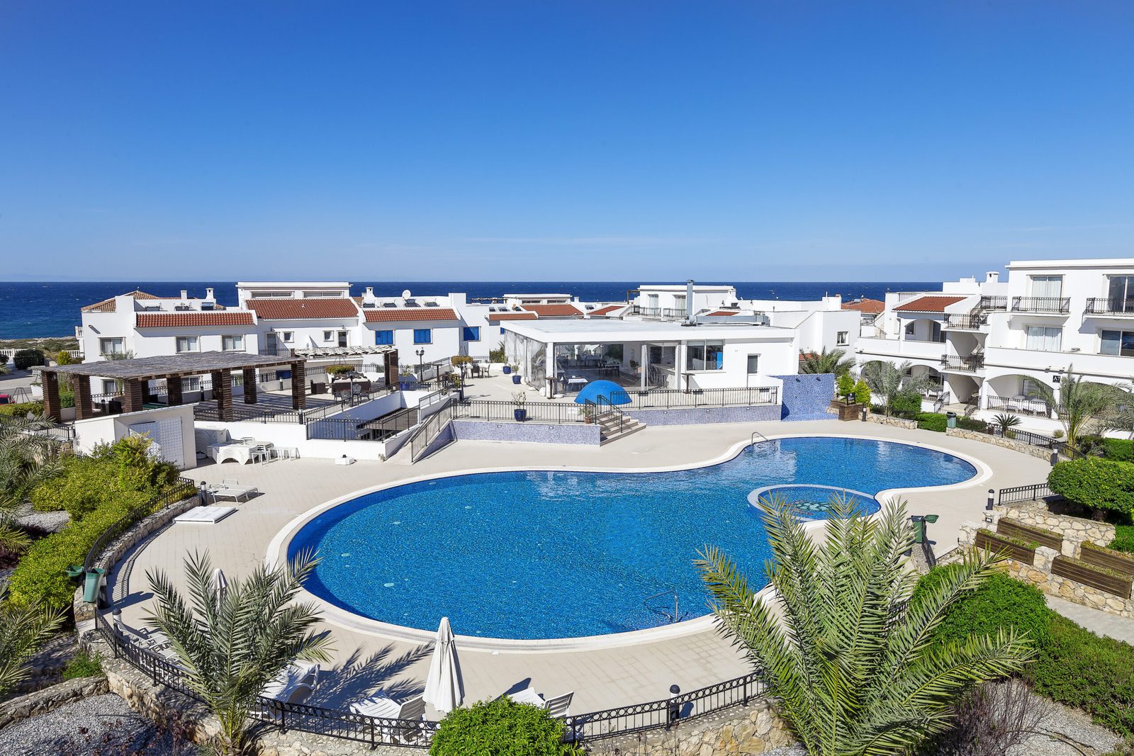 Elevated photo of a free-form pool, sun terrace and white buildings with the sea behind